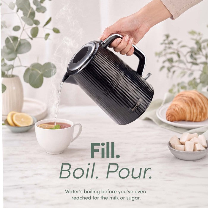 Hand pouring hot water from a black ribbed Keplin electric kettle into a cup of tea, with the kettle tilted above the cup.