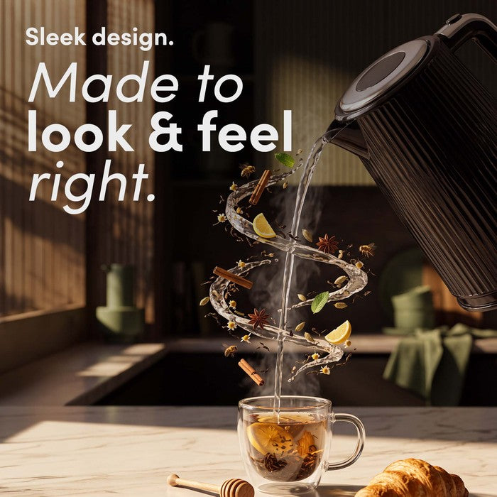 Black ribbed Keplin electric kettle pouring water into a glass cup of tea, with floating lemon, mint, spices and other tea ingredients around the stream.