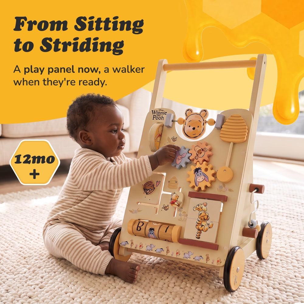 Baby sitting on a rug playing with the gears on the Winnie the Pooh wooden activity walker for 12 months+